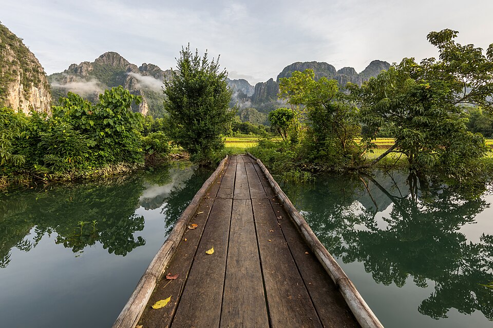 Front view of a wooden footbridge over a lagoon, trees and mountains in Vang Vieng, Laos